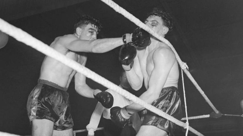 Boxer Jack Gardner punching Johnny Williams in the face, causing him to lean back on to the ropes of the boxing ring.