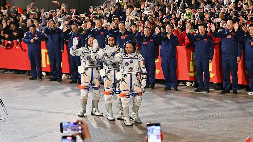 Commander Zhang Lu, Wu Fei and Zhang Hongzhang stand waving in space white space suits to a crowd of peple cheering them.