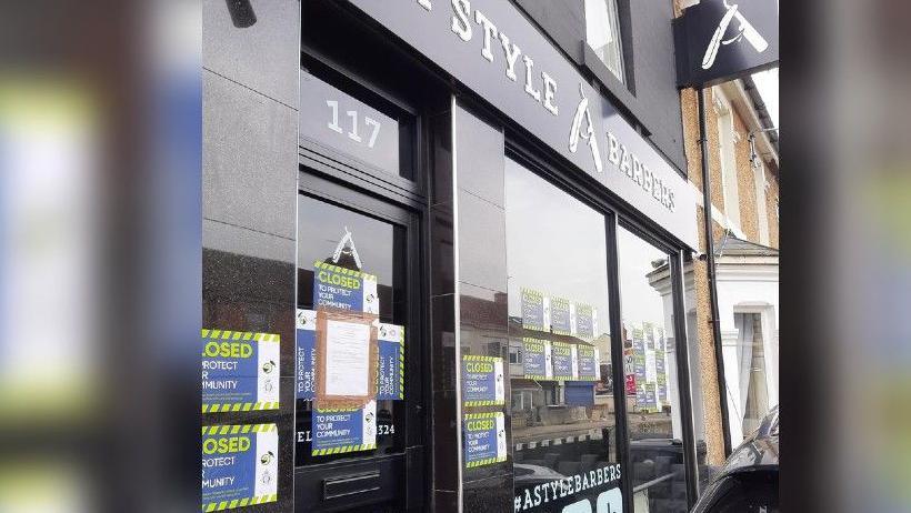 The exterior of the barbers in Swindon. It has around a dozen small 'closed' signs with police logos on the door and windows. The exterior is black and has large white lettering saying 'A Style Barbers'.