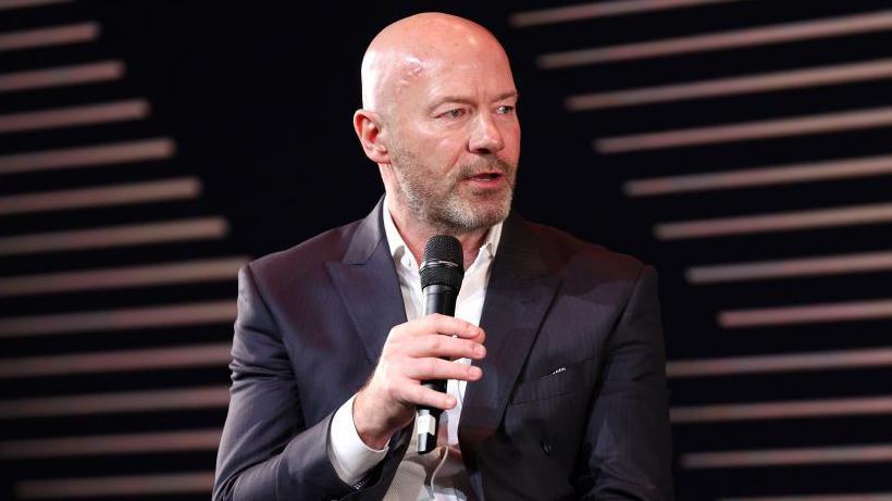 Alan Shearer wearing a black suit and white shirt, talking into a microphone