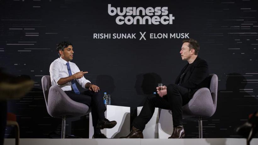 Rishi Sunak (a man with dark hair wearing a white shirt and blue tie) on stage interviewing Elon Musk (a man with dark hair in black clothing) at a "business connect" event