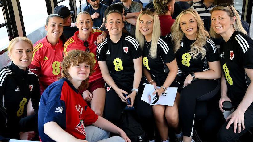 A group of women dressed in black and red football kits, all looking at the camera and smiling