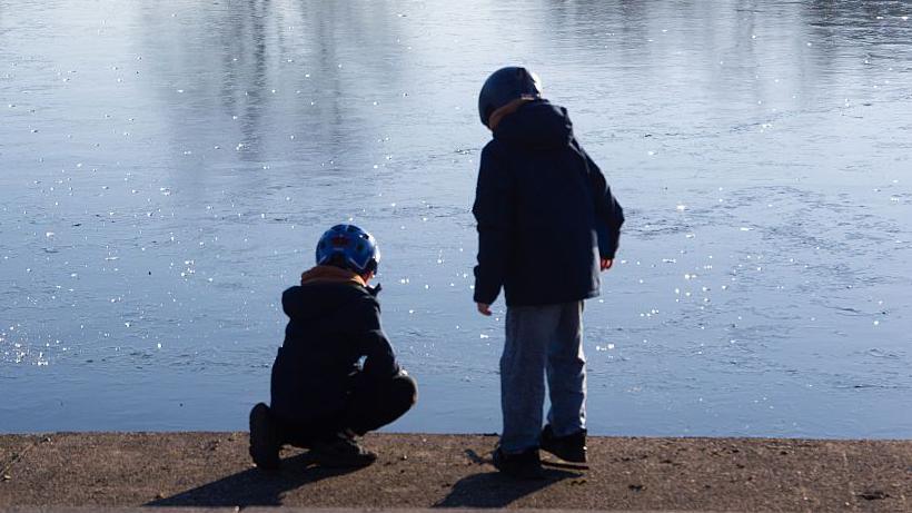 Two children peer at a frozen body of water in a park. 