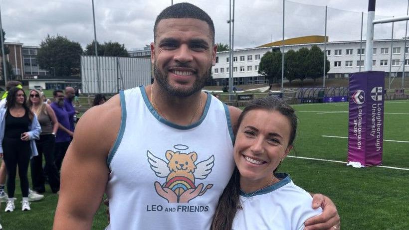Zack George wearing a white vest with a motif of a yellow teddy bear with wings and a halo, and a pair of hands opening to reveal a rainbow. He is standing with his arm around his wife Samantha who is wearing a white t-shirt. They are both smiling. 