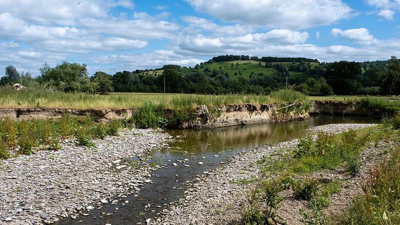 A mostly dried out river bed with some water in it in a landscape of green grass and hills in the background with a blue sky with white clouds above 