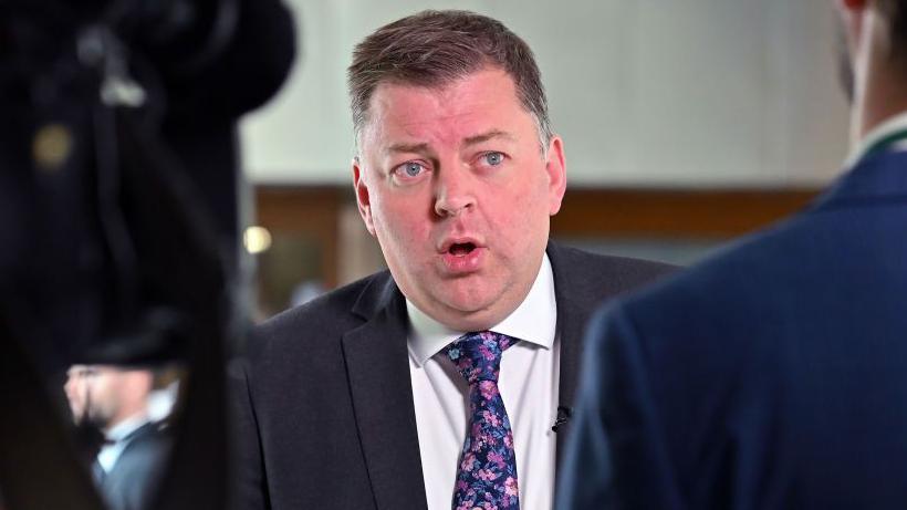Colin Smyth, a man with spiky brown hair mid speech while wearing a suit, white shirt and blue flowery tie