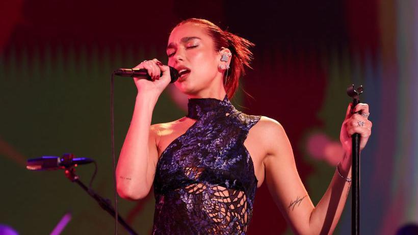 Woman in purple dress singing into microphone against a dark background