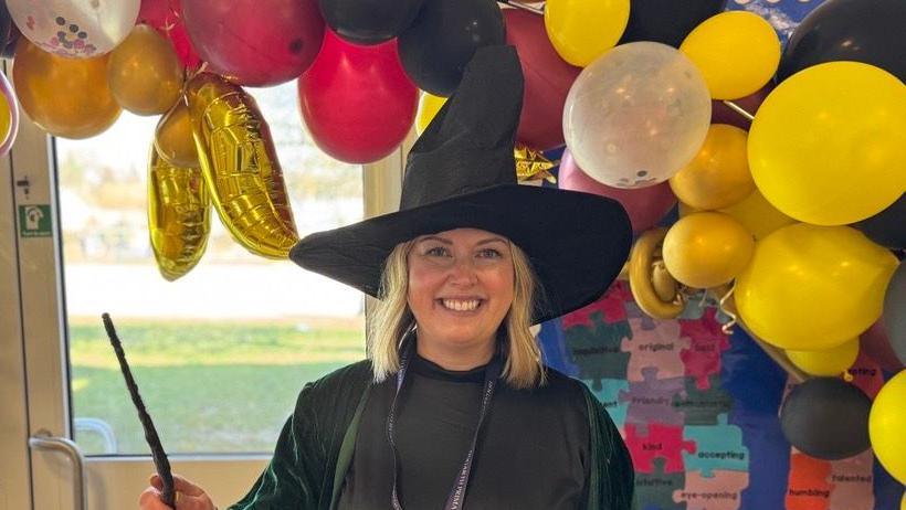 A woman with shoulder-length blonde hair wearing a green cloak, black dress, and witch's' hat while holding a wand. She is smiling straight at the camera and standing in front of a balloon arch.