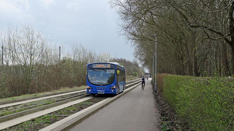 A blue bus travelling along the busway in 2018. A man is cycling next to the busway. The bus travels between straight concrete tracks.