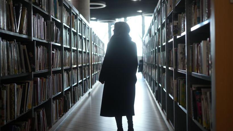 A woman can be seen walking along the aisle of a library in the dark.