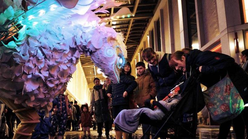 A person dressed in a large pale coloured bird puppet outfit is leaning over a child in a buggy with adults surrounding the buggy and looking on.