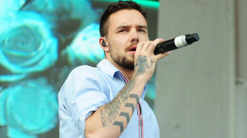 Liam Payne in a blue shirt holding a microphone up to his mouth with a stage background on blue flowers behind him