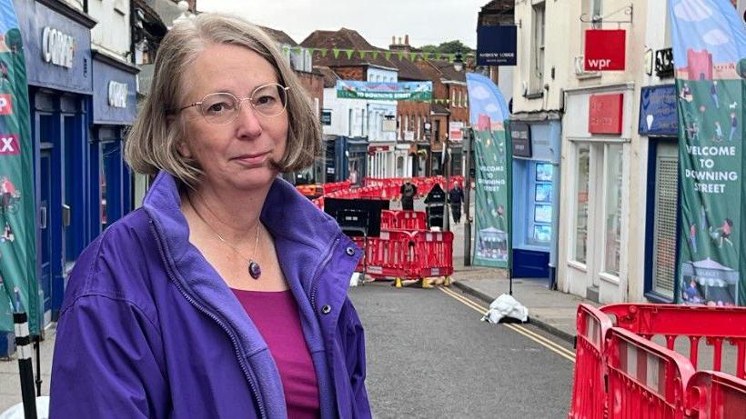 A middle aged caucasian woman with short bobbed brown hair and glasses smiles slightly at the camera wearing a purple jacket with roadworks along the high street behind her.