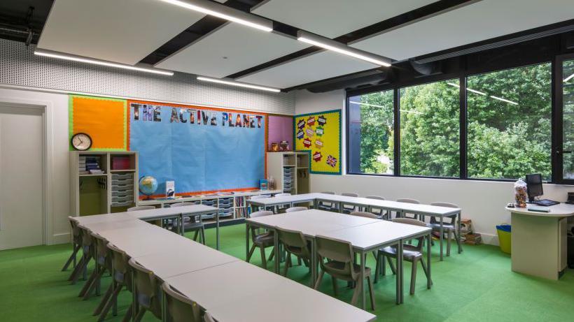 A stock photo of the inside of a primary school classroom. There are tables with numerous chairs and a teacher's desk.