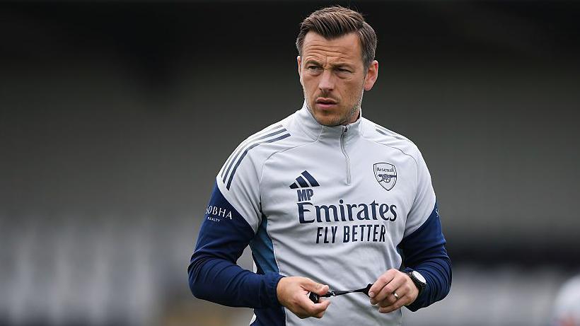 Max Porter takes a training session at Arsenal. He is wearing a grey track top with navy blue sleeves and is stretching the chord of his whistle between his hands as he watches on.