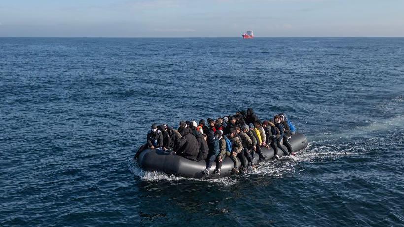 A black inflatable boat in a large body of water with a crowded group on it. 