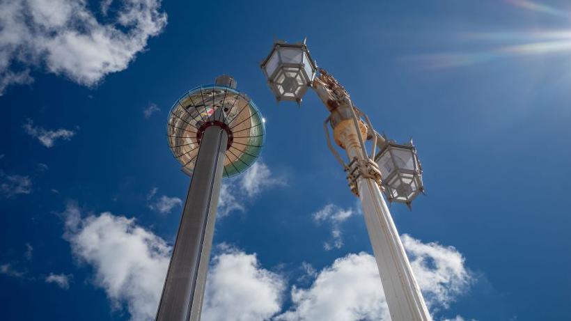 The i360 viewing tower in Brighton, pictured from the ground. The image has blue skies with some clouds and two lampposts to the right of the i360.