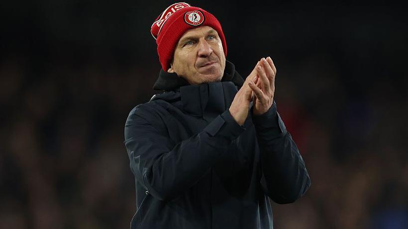Gerhard Struber wearing a red Bristol City beanie hat and dark overcoat applauds supporters