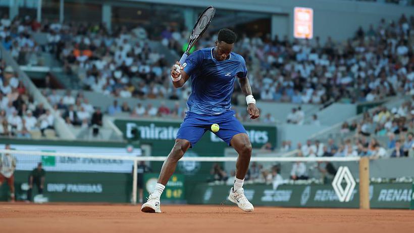 Gael Monfils, wearing a blue T-shirt and shorts, lines up a shot between his legs with the ball between his thighs and his black racquet held in his right hand at head height