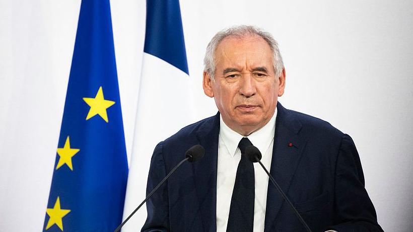 The French prime minister wears a dark jacket and tie and stands in front of a French and European Union flag