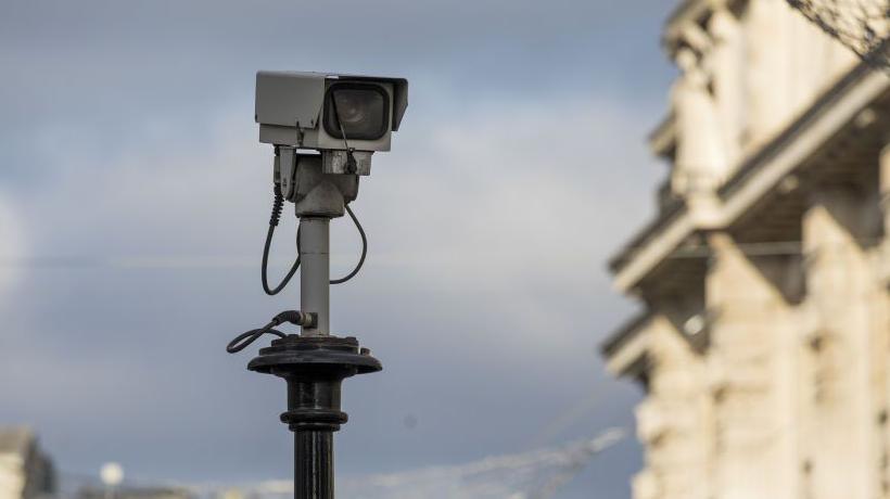 A stock image of a grey CCTV camera is on top a black pole. In the background there is blue sky and an out of focus building. 