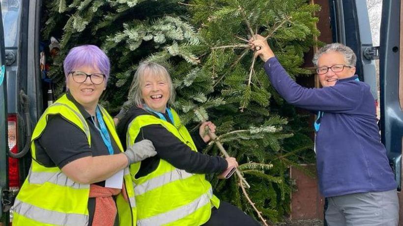 Three older women smile and laugh as they hold onto Christmas trees that have been loaded into the back of a van. Two of them are wearing hi-vis waistcoats.