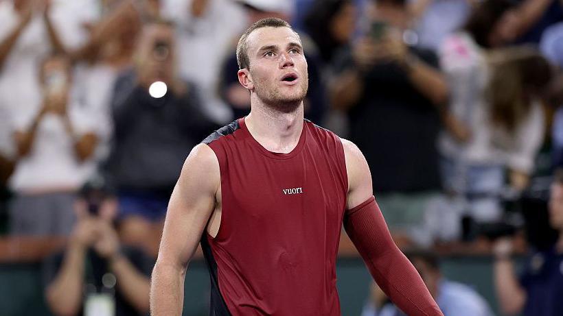 Jack Draper, wearing a maroon vest with a maroon sleeve on his left arm, looks upwards with his mouth open 