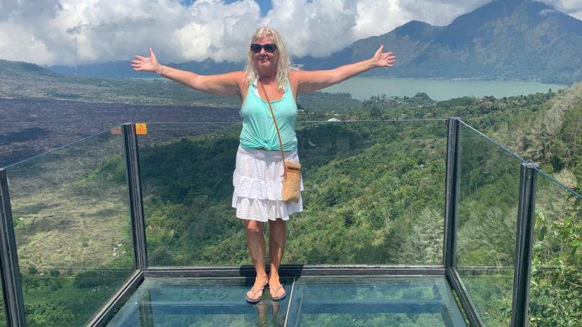 Nicky Maidment stands with her arms outstretched on a glass balcony overlooking some mountains. She wears a light blue tank top and a white frilly skirt.