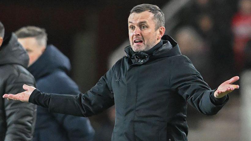Nathan Jones extends his arms outwards, wearing an overcoat, on the touchline at a Charlton Athletic match