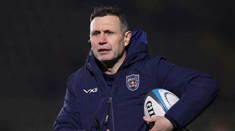Stephen Jones holding a rugby ball under his left arm and with an earpiece in his right ear, warming players up for a Newcastle game 