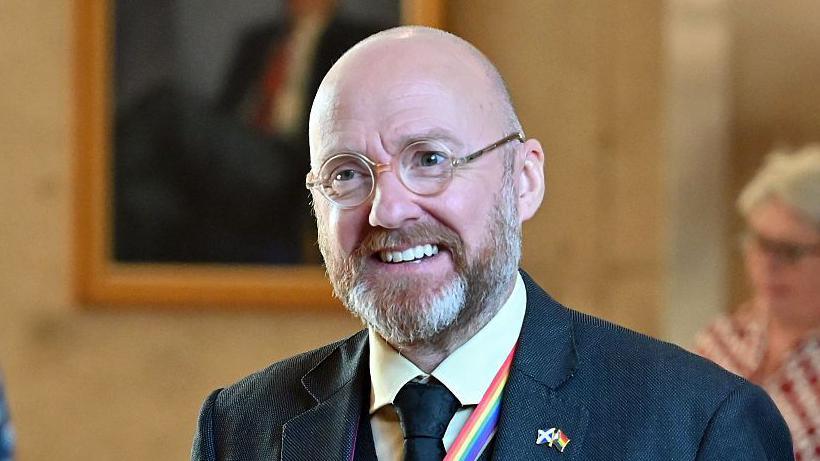 Patrick Harvie is a bald man with round glasses and a grey beard. He is wearing a blue suit, yellow shirt and blue tie