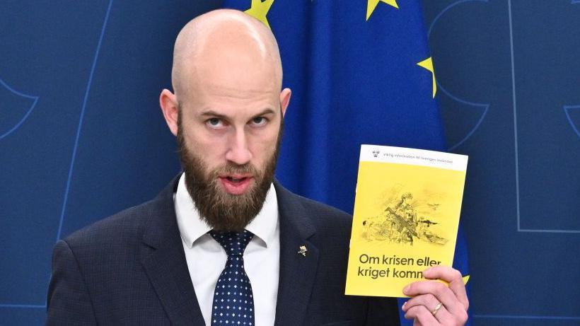 Sweden's Minister for Civil Defence Carl-Oskar Bohlin holds a copy of  the new version of the preparedness booklet "If the crisis or war comes". The pamphlet has a yellow cover and an image of a woman in army fatigues holding a large gun.