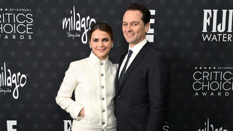 Matthew Rhys with Keri Russell. Matthew is wearing a black suit and tie while Keri is wearing a white tweed coat. They stand in front of a black background with white logos. 