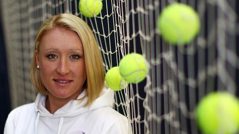 Elena Baltacha smiles at the camera while standing next to netting which has numerous tennis balls stuck in it. She has blonde hair cut in a bob style and she is wearing a white hoodie with pearl earrings.