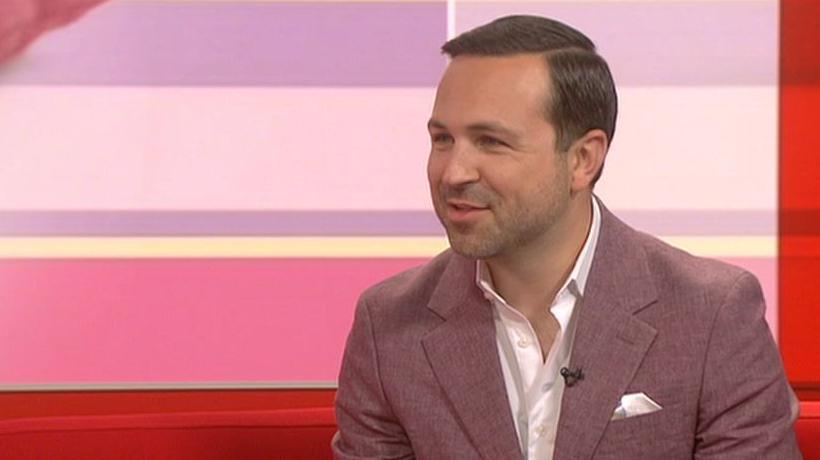 A man with short dark hair, a white shirt and a pink jacket in a studio with a red and pink background
