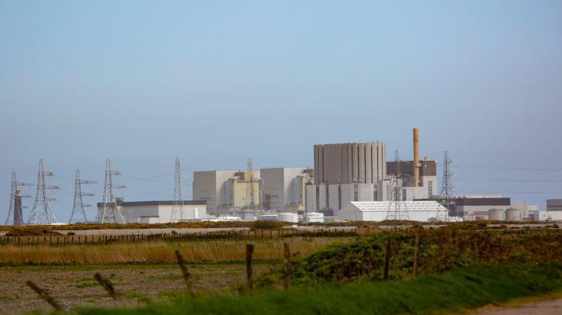 Dungeness B power plant. It is a series of large grey buildings, pylons and one tall chimney. In the foreground there is a grassy field.