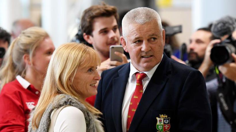 Gatland and his wife facing eachother while a a group of fans stand around them. They are looking off to the right and smiling.
