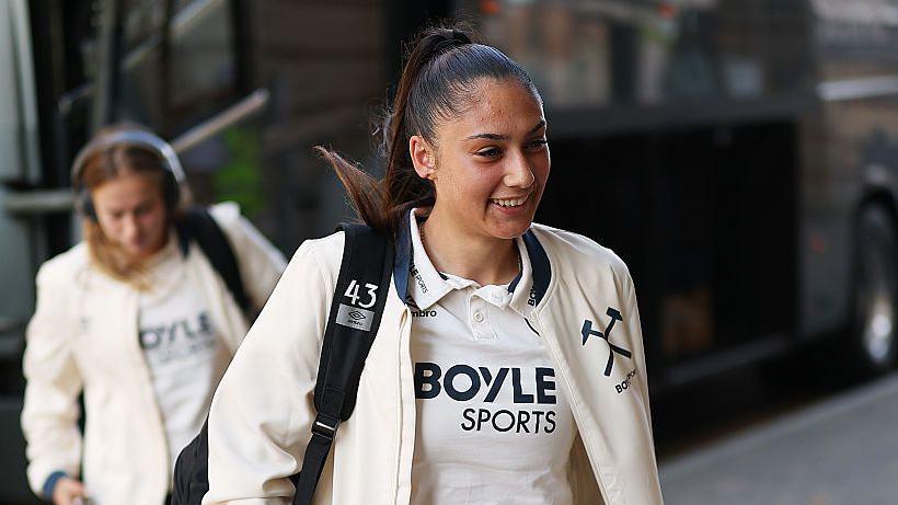 Selin Cemal steps off the team coach as West Ham prepare to face Tottenham in September 2025. She has dark hair, tied back, and is wearing a wearing jacket and white shirt with a Boyle Sports sponsor logo on the front. One of her team-mates is in the background and has a pair of headphones on.