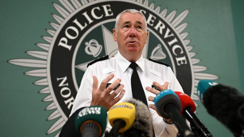 Chief Constable Jon Boutcher stands in front of several media microphones wearing a white shirt with black PSNI badges and a black tie. A large PSNI logo looms over him in the background.