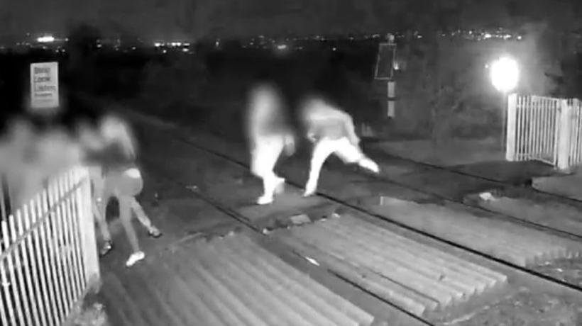 Two people running across a railway track in the dark, with two other people at a closed gate at the edge of the track. All faces are blurred.