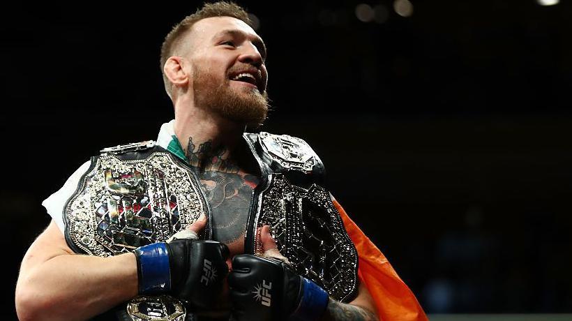 Conor McGregor smiles sitting on top of the cage with both his UFC belts around his shoulders