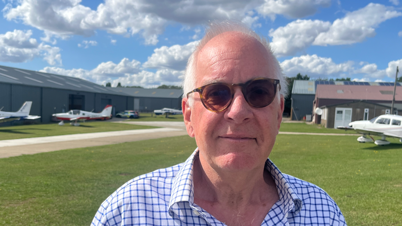 A head and shoulders image of Charles Course, who is the owner of Elmsett Airfield. He is wearing sunglasses and a shirt. He is looking into the camera. Planes can be seen in the background.