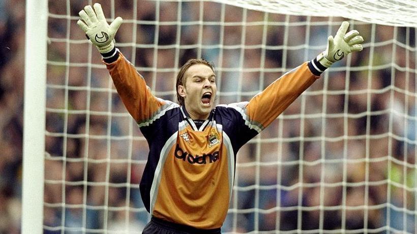 Manchester City goalkeeper Nicky Weaver, wearing an orange jersey with black sleeves, has his arms raised above his head after saving a penalty.