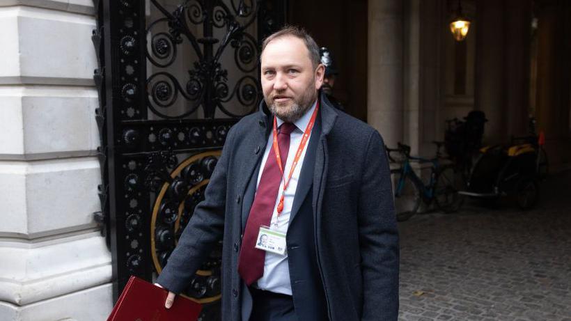 Ian Murray - a man with light coloured hair and a beard, wearing a dark jacket and a red tie - walking out of a government building in London, holding a red folder