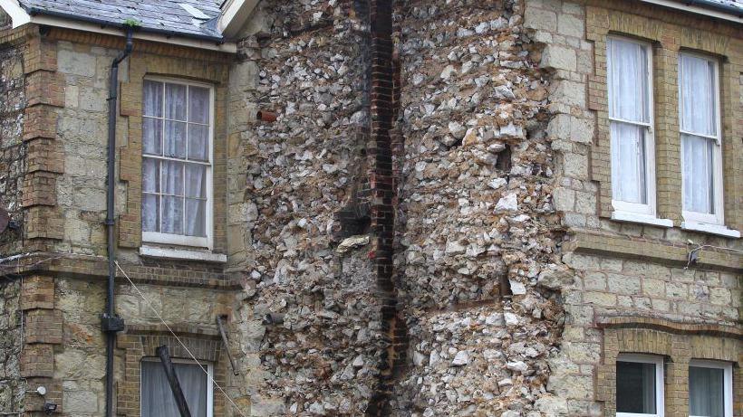 Side view of pale brick and stone house with white windows.  A large section of a wall has collapsed onto a car parked below.