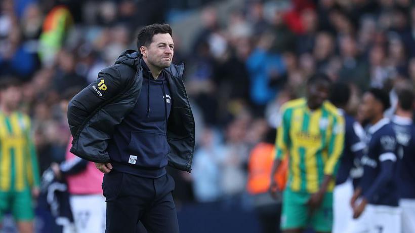 West Brom manager Ryan Mason adjusts the back of his sweatshirt and stares thoughtfully into middle distance after defeat