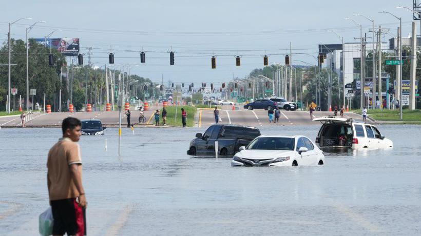 A young man wades through flood waters that come up to the top of car wheels