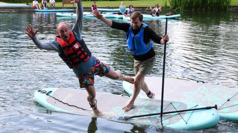 Ed Davey falling off a surfboard and into the River Thames, as Freddie van Mierlo gets set to also fall into the water 