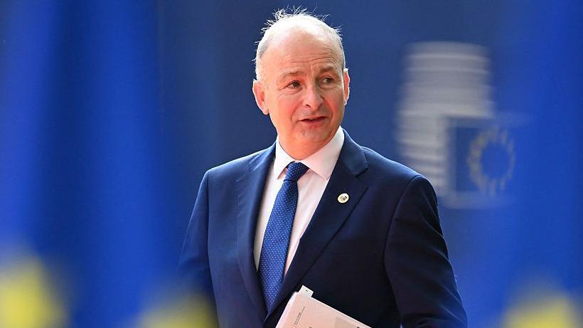 Martin wearing a navy suit jacket, blue tie and white shirt. He is looking to the side and speaking. The background is mainly bright blue.