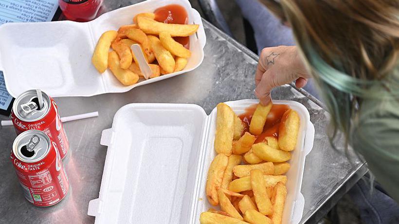 Close up cartons of chips and fizzy drinks and a hand holding a chip and dunking it in ketchup 